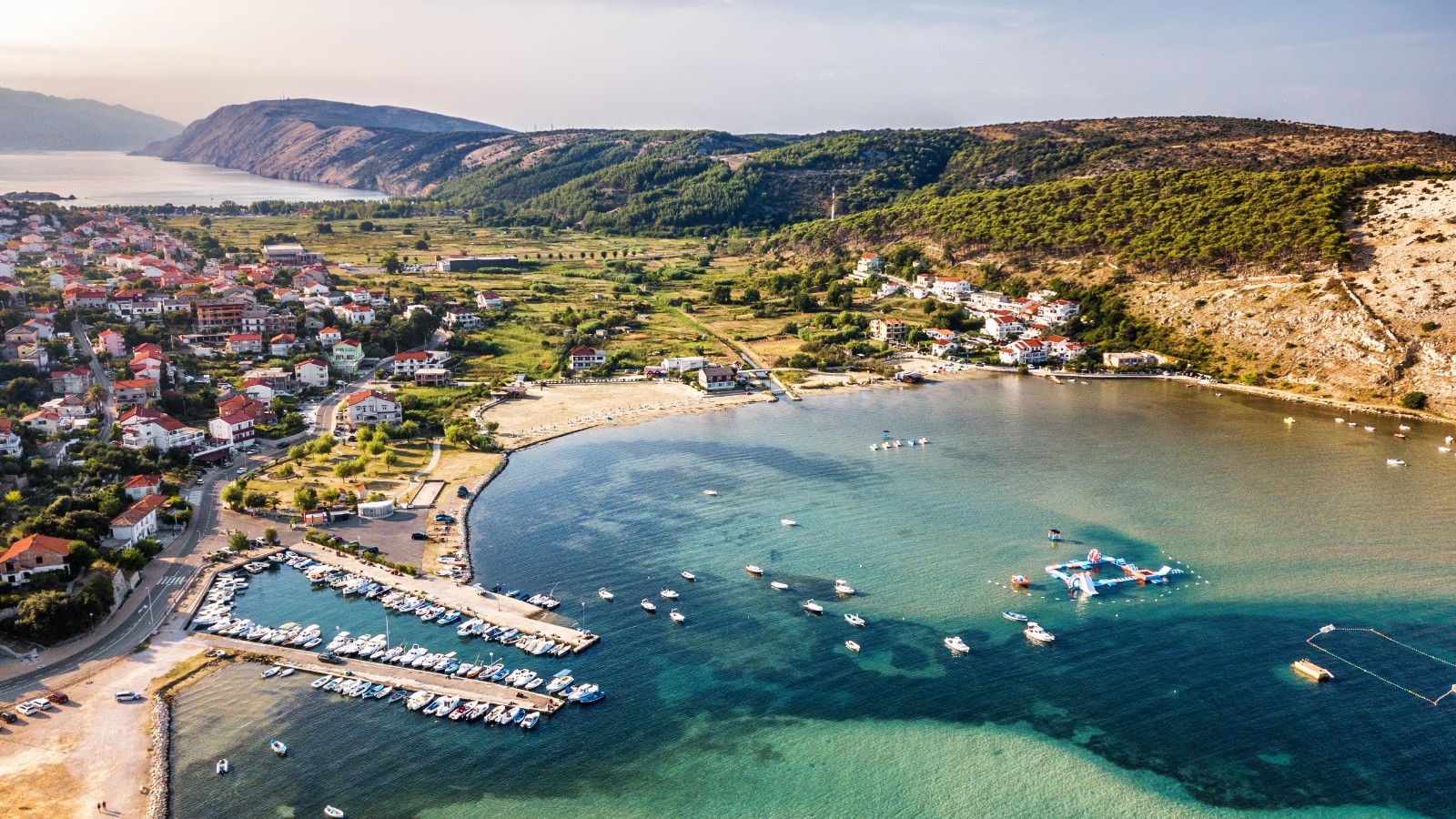 Aerial view of Lopar bay on the island of Rab — a marina full of small boats, a sandy beach and clear turquoise Adriatic water backed by green hills — Croatian National Tourist Board / Aleksandar Gospić